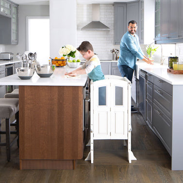 A man and a boy wash their hands at a kitchen counter, with a wooden step stool beside the boy.