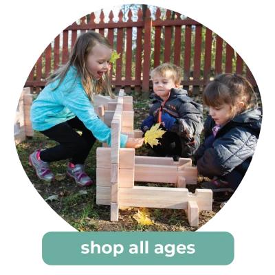 Three young children play with wooden blocks outdoors near a red fence. Shop toys for all ages.