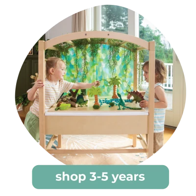 Two young children play with dinosaur toys at a wooden activity table designed for curious minds. Text reads shop 3-5 years.