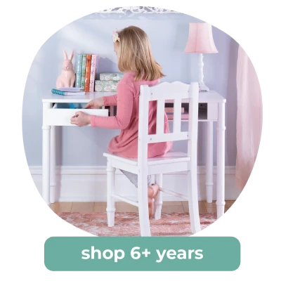 Young girl sits at a white desk, opening a drawer, surrounded by books and decor. Perfect spot for creativity. Shop 6+ years.