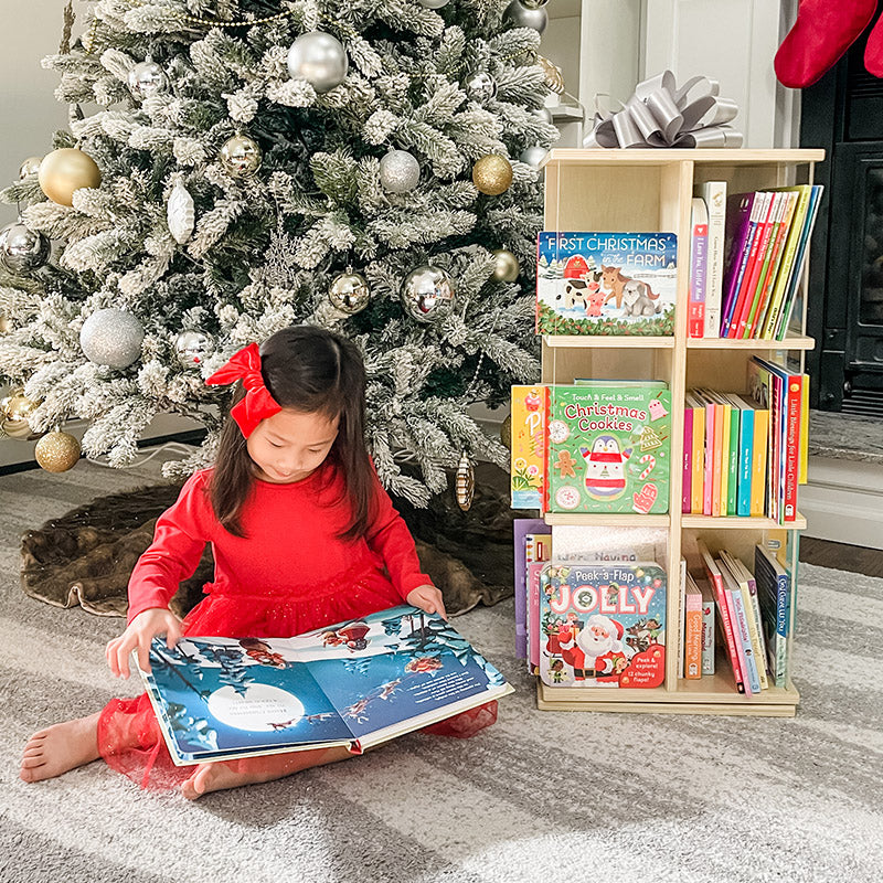 Young girl reading a holiday picture book near a natural wood Guidecraft Rotating Book Display