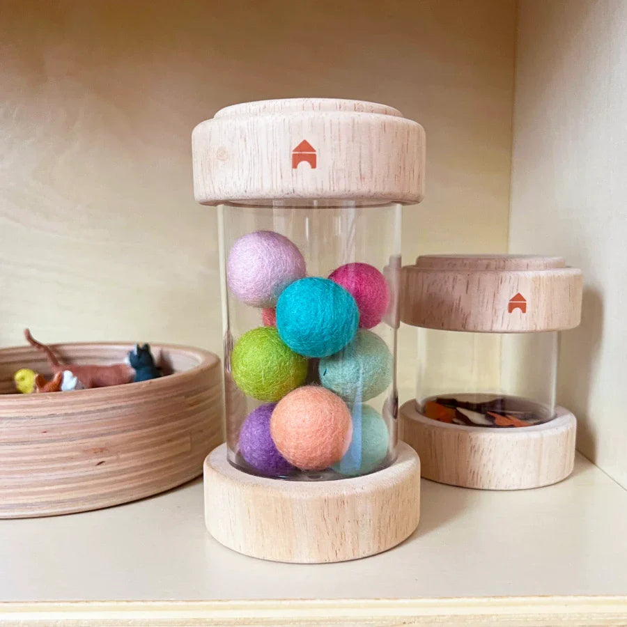 A Guidecraft clear Treasure Tube filled with colorful felted wool balls, surrounded by other containers holding assorted interesting small play materials
