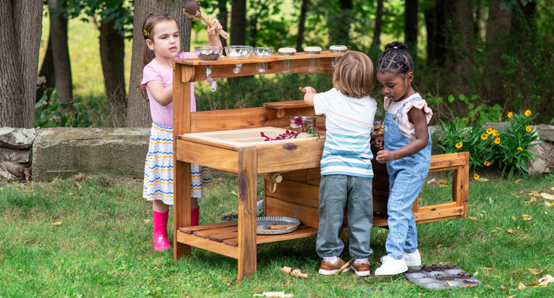 Three young children play together at a wooden outdoor mud kitchen in a grassy, wooded area.