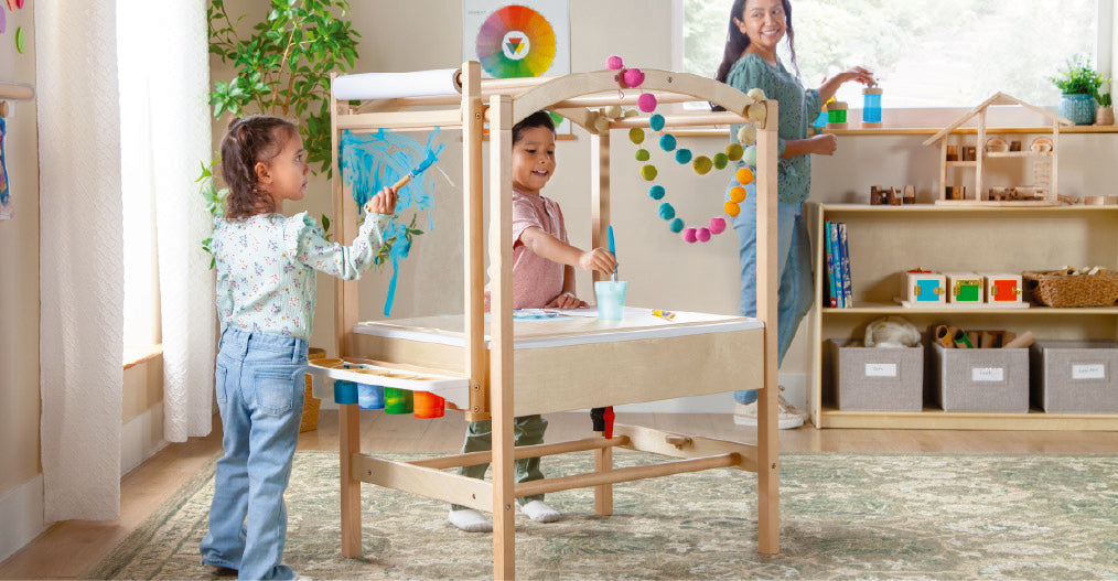 Two young children draw together on a chalkboard easel in a bright, modern playroom.