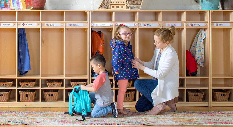 A teacher helping a young preschool child put her coat on in front of a line of wooden lockers while another fills his bookbag nearby