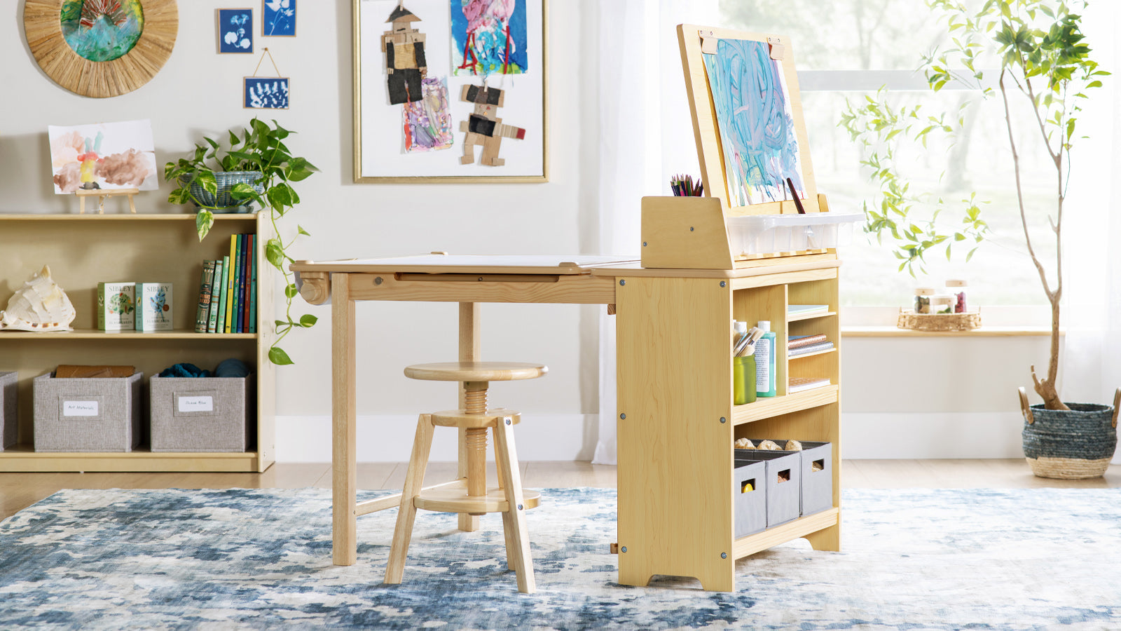 A wooden art desk with supplies, stool, and artwork, set in a bright, cozy room with plants and shelves.
