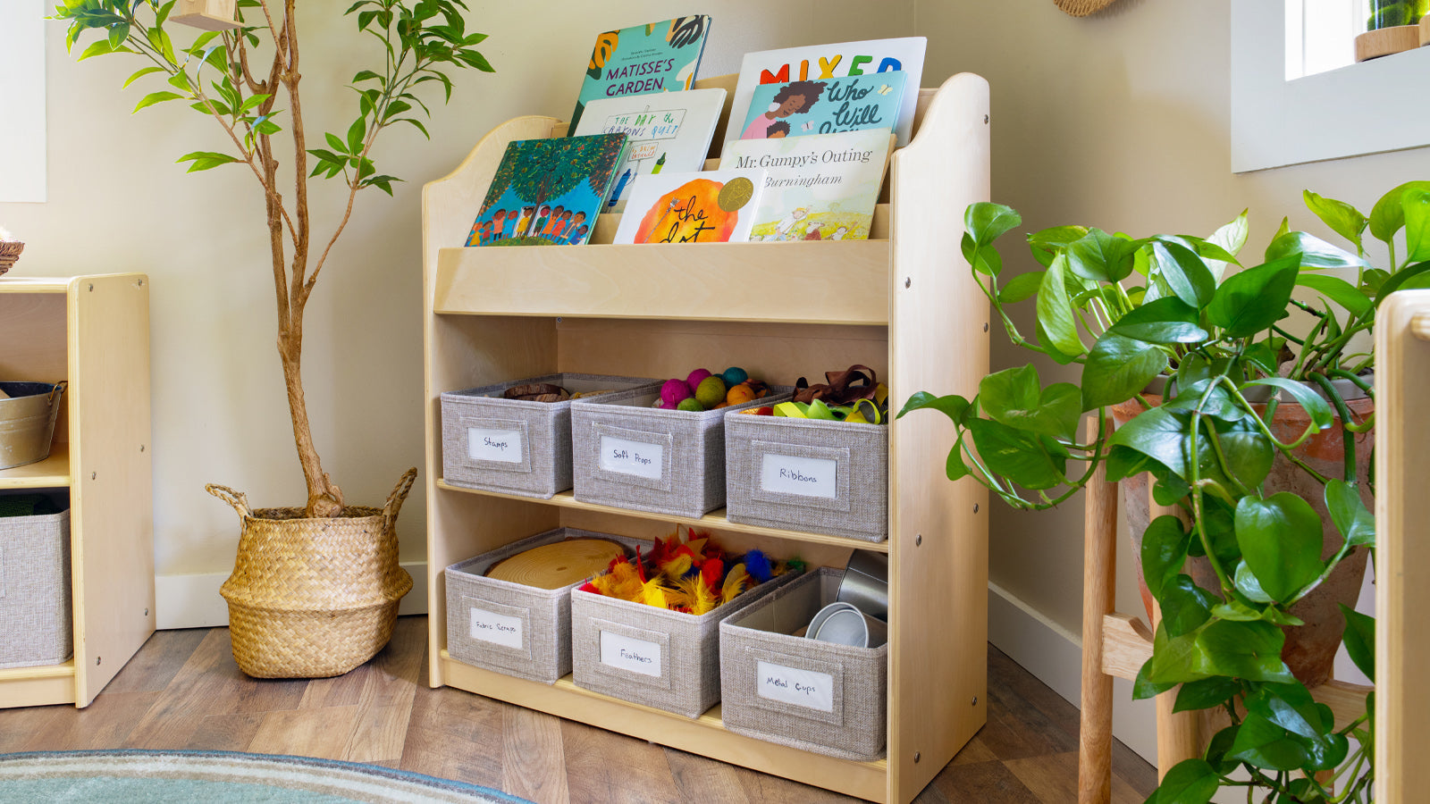 A wooden shelf holds children’s books on top and labeled fabric bins with toys and supplies on the lower shelves.