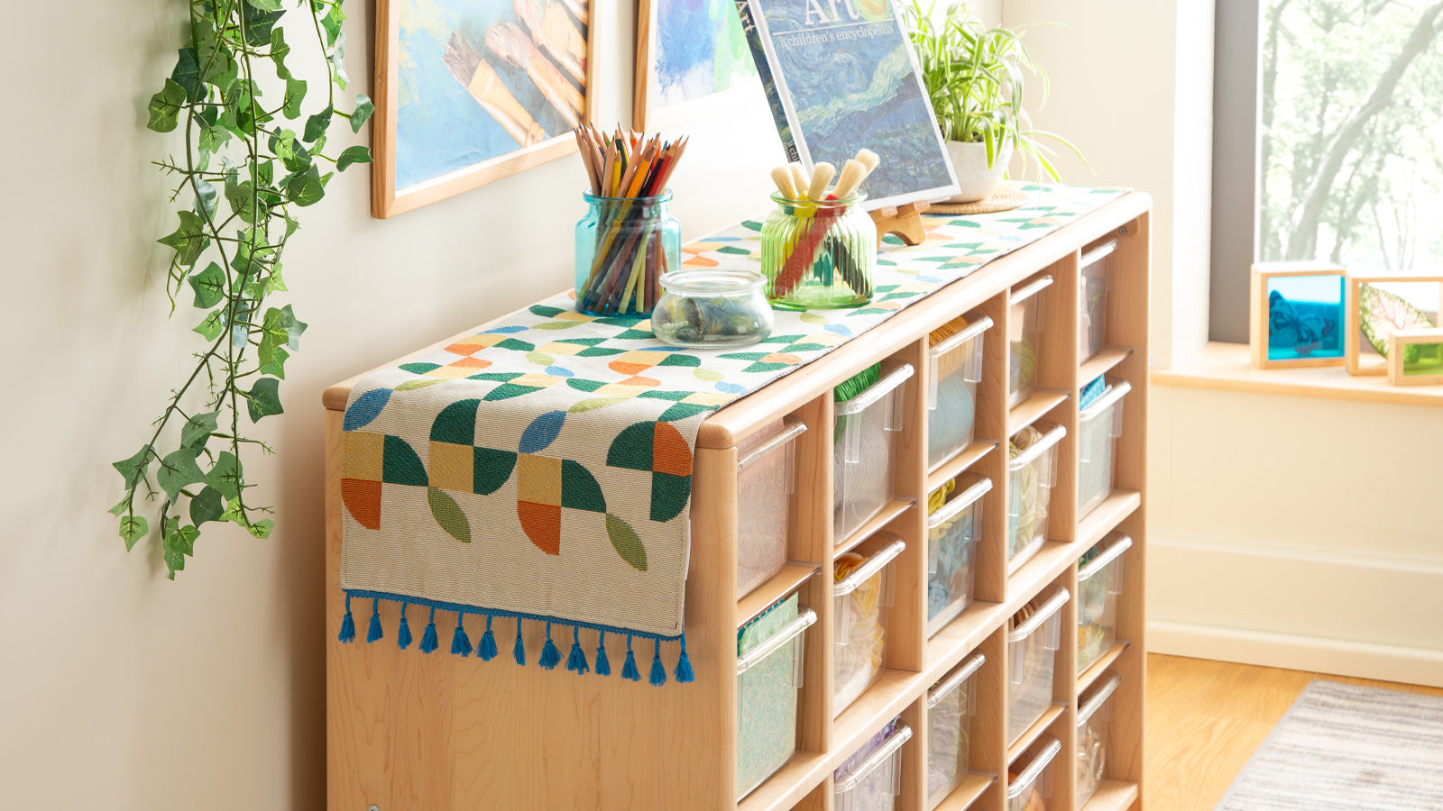 Wooden shelf with clear bins, art supplies, and decor, next to a window with natural light and green plants.