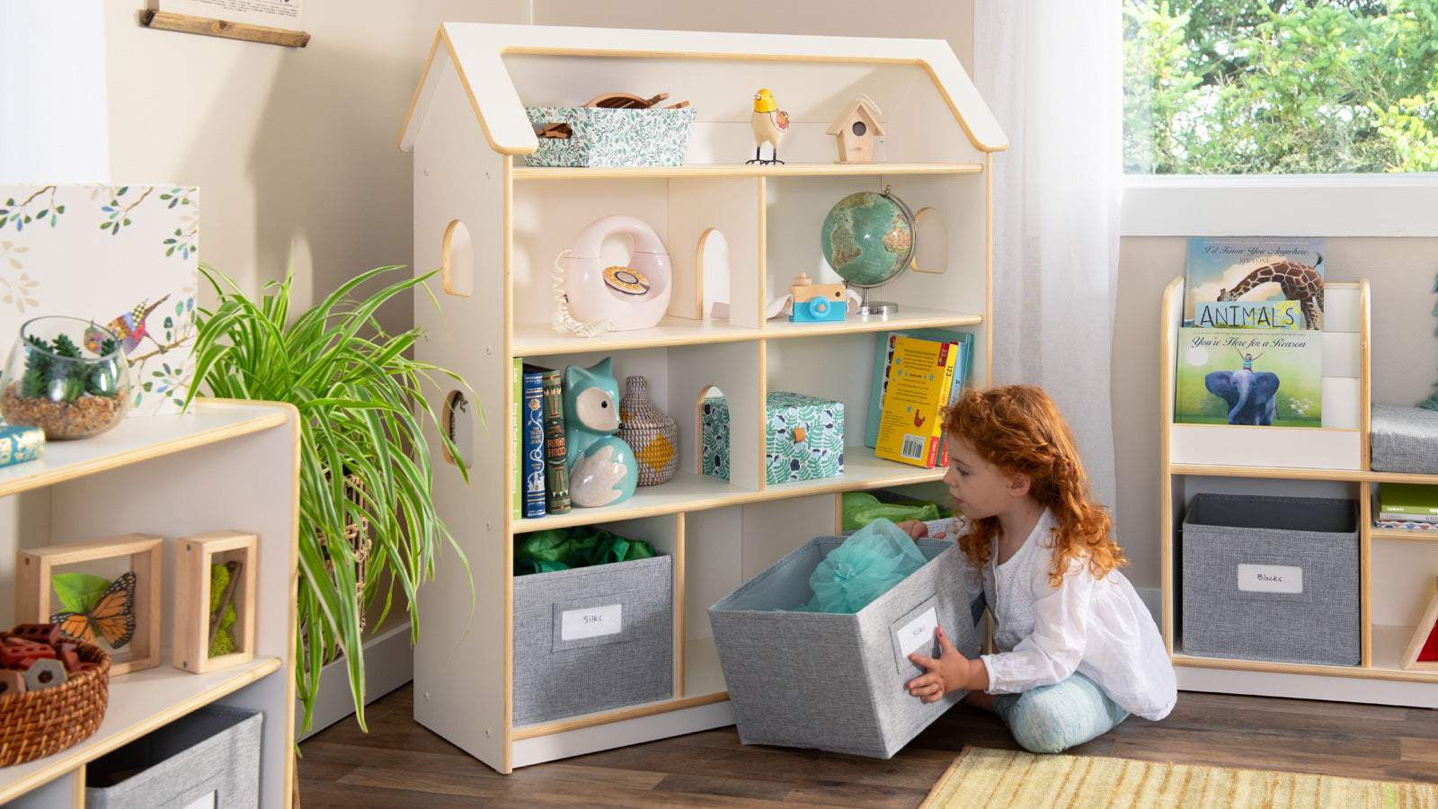 A young girl kneels by a toy shelf, pulling out a gray storage bin in a bright, organized playroom.