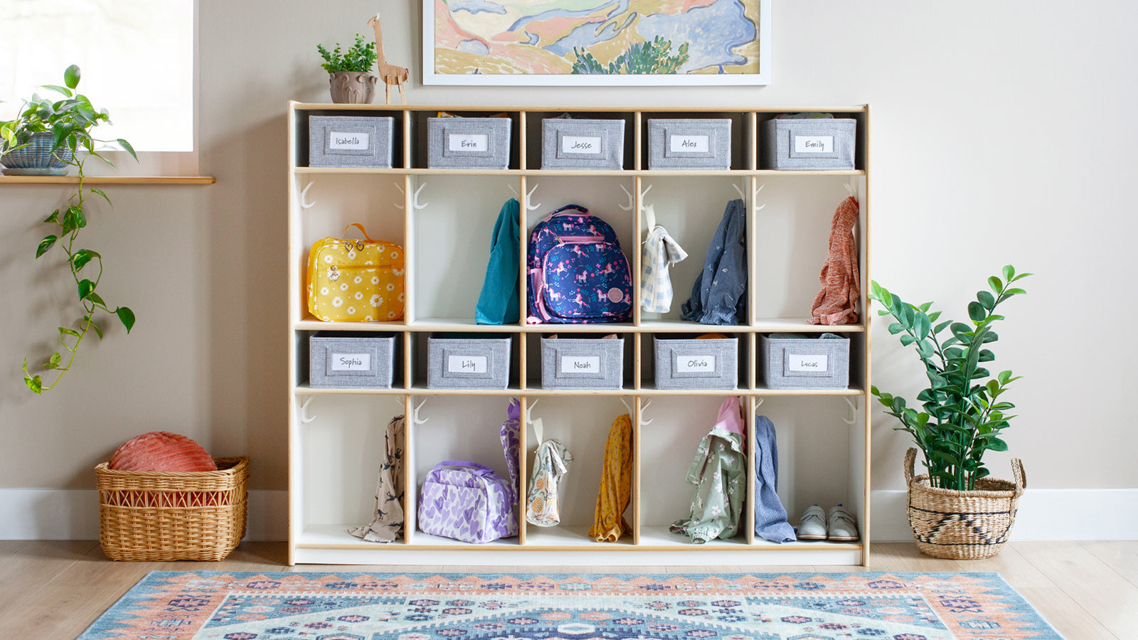 A cubby storage unit holds backpacks and jackets, with name labels, in a bright, organized classroom.