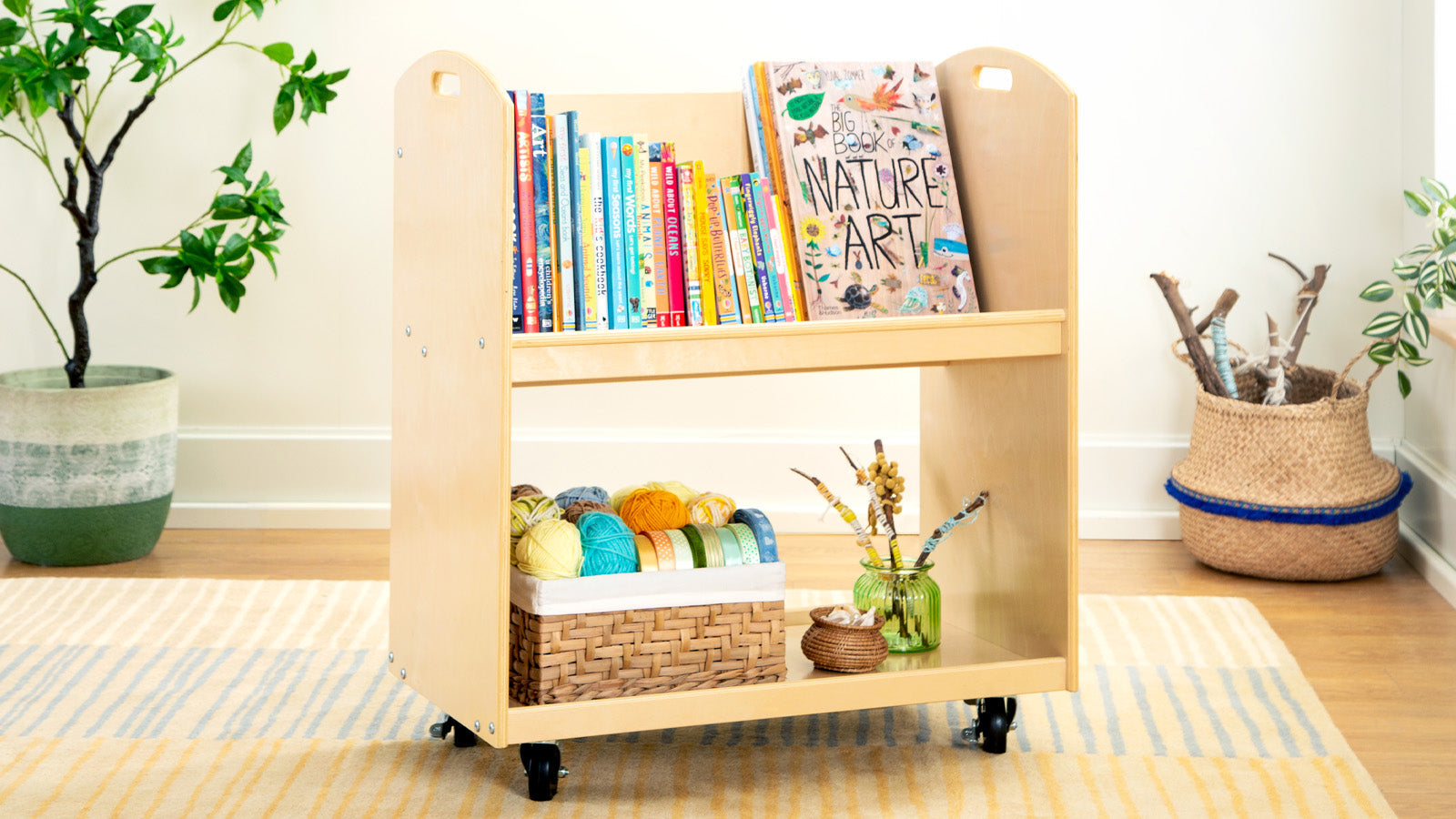 Wooden book cart with childrens books on top and yarn, a basket, and decor on the bottom shelf in a cozy room.