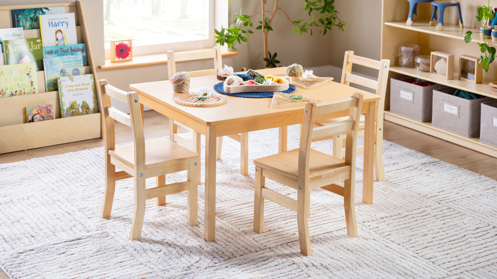 A small wooden table with four chairs sits on a rug in a bright, cozy classroom with books and plants.
