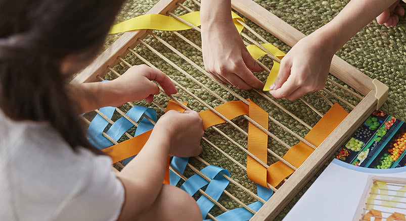 Two preschool-aged children weaving multicolored ribbons onto a weaving frame matrix