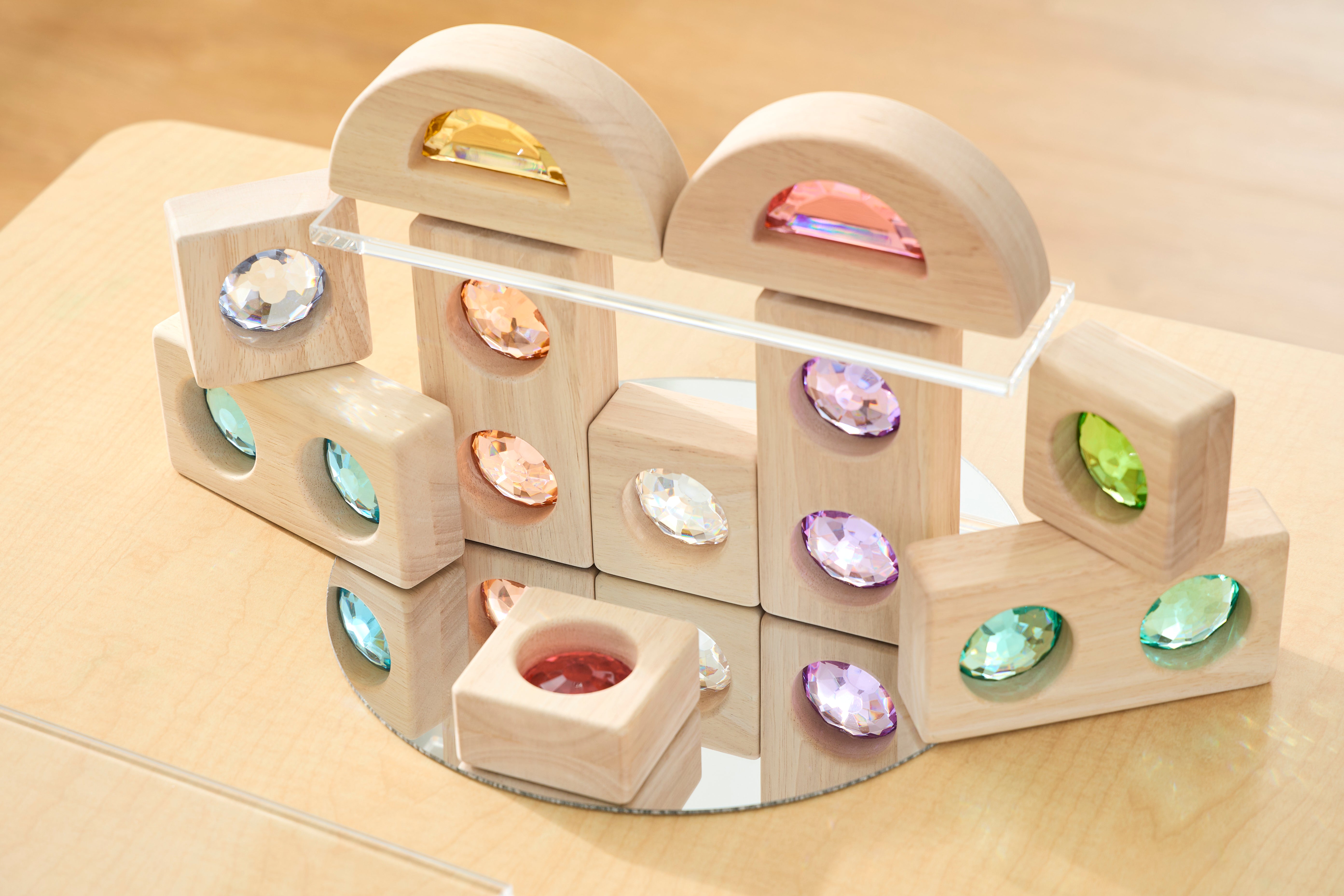 Wooden blocks with colorful gem-like inserts are arranged on a mirror atop a light wooden surface.