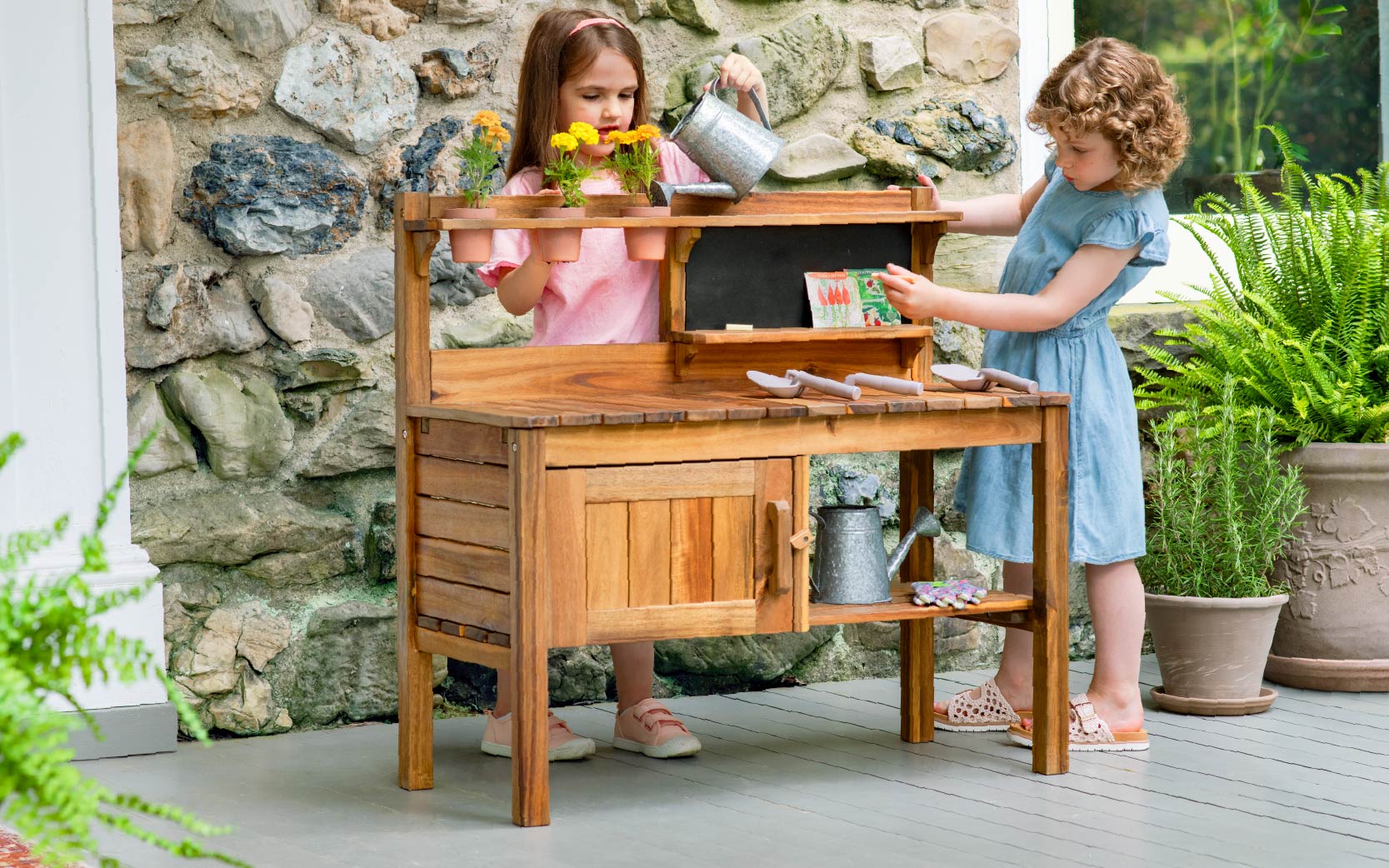 Two young girls play with a wooden potting bench, watering and arranging flowers outdoors on a porch.