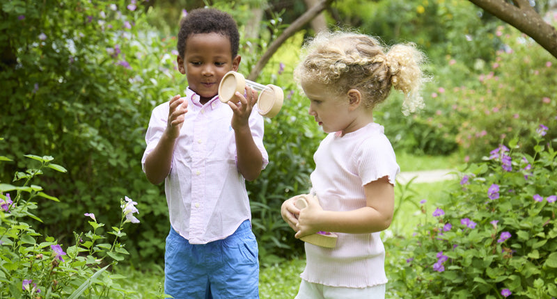 Two young children play with wooden cups in a garden surrounded by green plants and purple flowers.