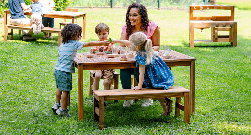 An outdoor classroom scene with several teachers and preschool-aged children working at a table with natural loose parts and Guidecraft treasure tubes. An outdoor mud kitchen is visible in the background