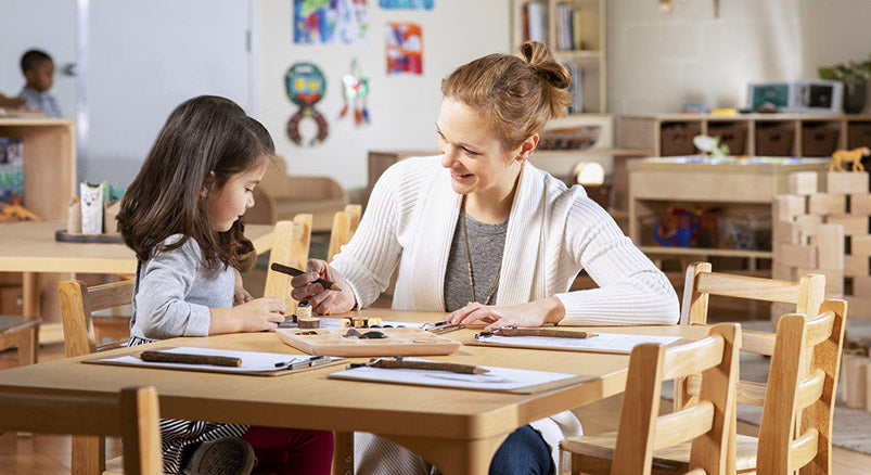 A young female teacher working on writing skills with a young female preschool aged student at a wooden table surrounded with wooden chairs