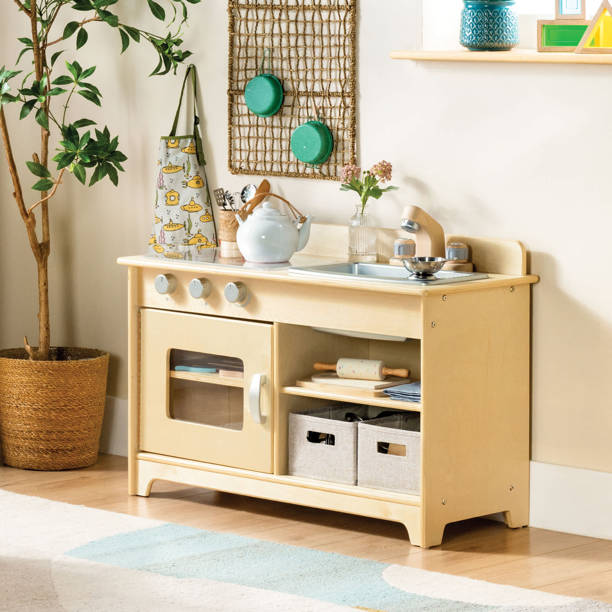 Wooden play kitchen with toy stove, oven, sink, and shelves, next to a plant and wall decor.