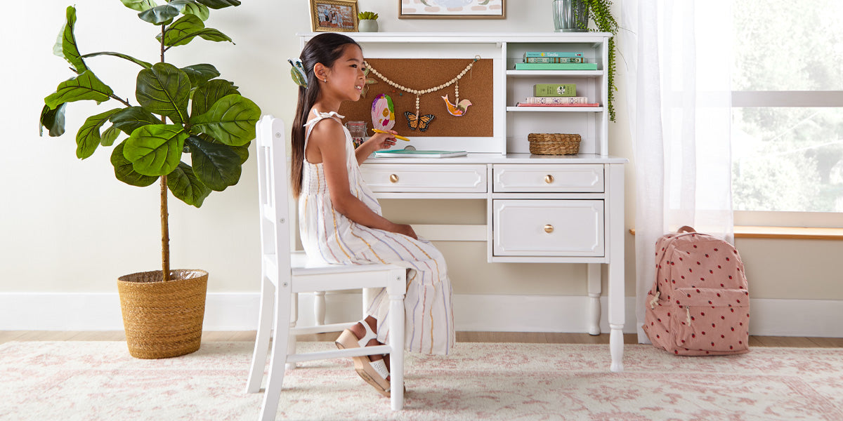Young girl sitting at a white desk in a bright room with plants and a backpack.