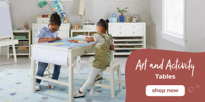 Two children draw at a white art table in a bright, colorful playroom. Text: Shop our art and activity tables now.