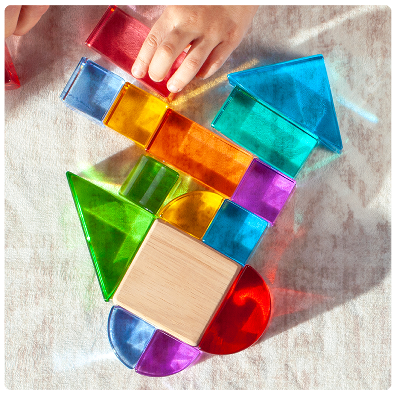 A close up detail image of a single, central square wooden unit block surrounded by smaller, mathematically aligned acrylic blocks that a child's hands are arranging next to the wooden block to show how several smaller pieces "add up" to the size of the larger pieces
