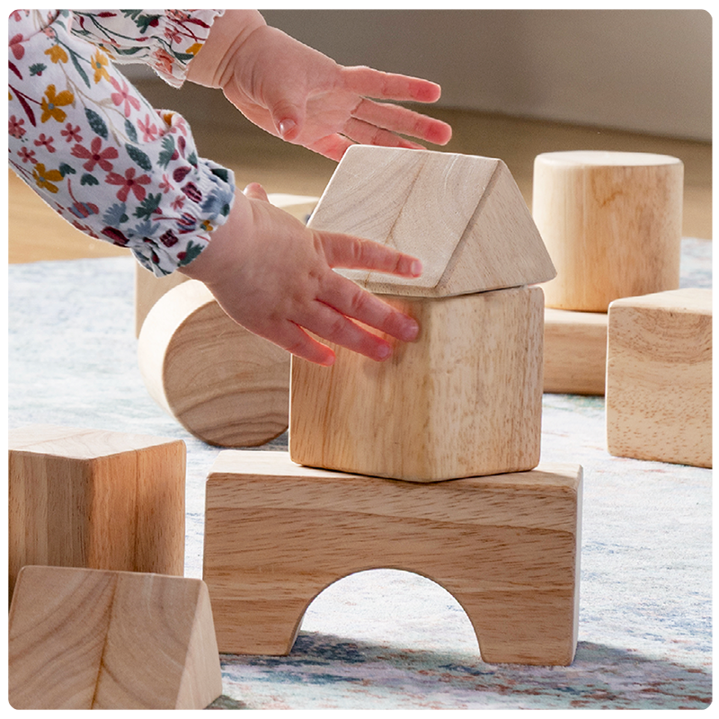 Close up detail image of a toddler stacking toddler unit blocks to test how they stack and balance. The hands are making a slight adjustment to the stacked blocks so they don't fall down.