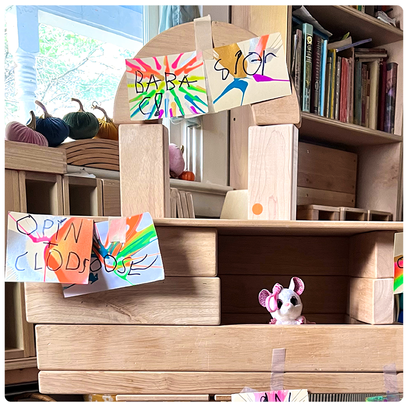 A child's unit block build that incorporates a child's handwritten and painted signage used to label the structure. There is also the use of other toys in this build like a small stuffed mouse as a charater in this child's built "store" structure