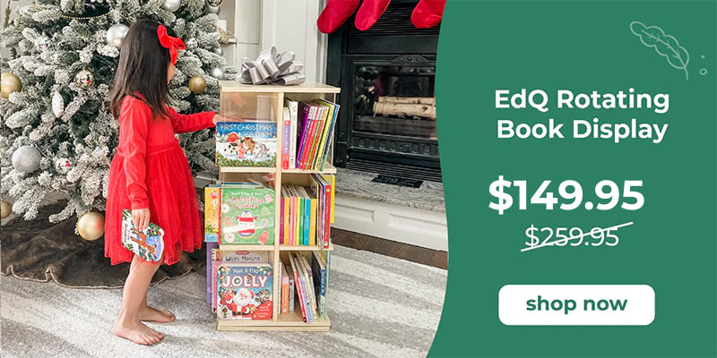 Child in a red dress standing next to a book display with Christmas decorations and a fireplace.