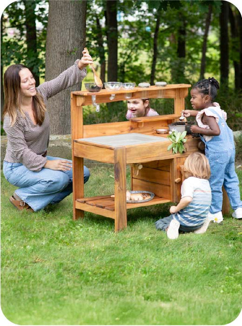 Woman and children playing with a wooden outdoor play kitchen set in a park.