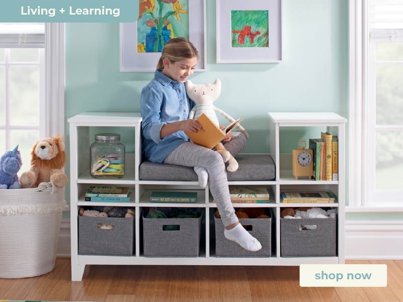 Child sitting on a white storage bench reading a book in a room with colorful artwork and toys. Click to shop the Martha tewart Living + Learning furniture collection.