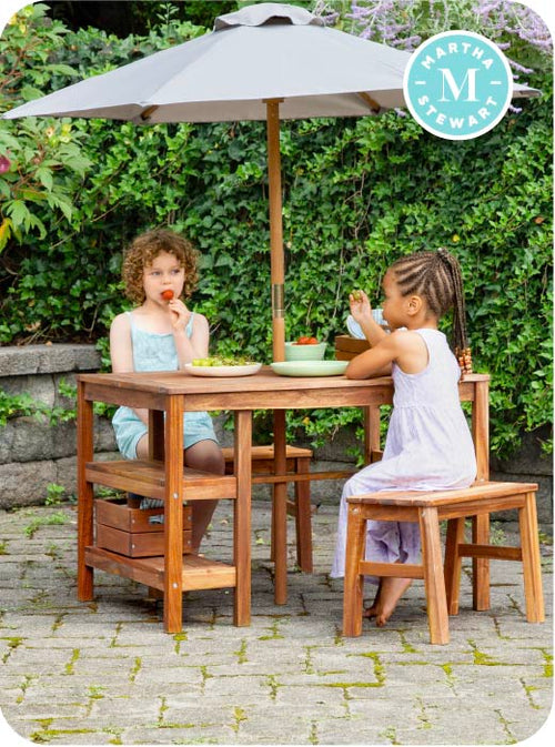 Two children sitting at a wooden outdoor table with a gray umbrella, surrounded by greenery.