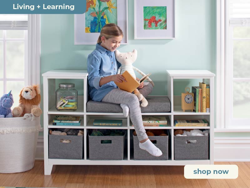 The child is sitting on a white storage bench, reading a book in a room with colorful artwork and toys. Click to shop Martha Stewart Living and Learning Furniture Collection