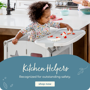 A toddler stands in a white kitchen helper high chair at a kitchen island, reaching for strawberries on the tray. A blue graphic overlay at the bottom reads 'Kitchen Helpers' with a 'shop now' button.