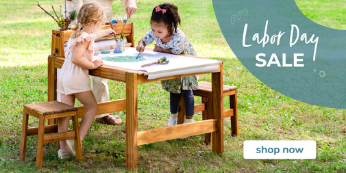 A wooden children's painting table and stools shown outdoors on grass with two young girls painting at the table. A large teal graphic in the upper right says 'Labor Day SALE' with a 'shop now' button below it.