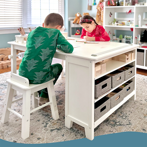 Two children sitting at a Guidecraft white desk with storage bins in a room with shelves and toys.