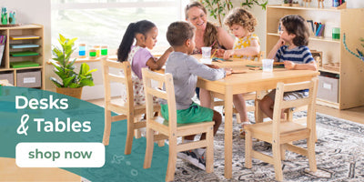 Children sitting at a table with a teacher in a classroom setting, featuring desks and tables.