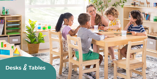 Children sitting at a table with a teacher in a classroom setting, featuring desks and tables.