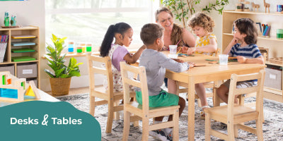 Children sitting at a table with a teacher in a classroom setting, featuring desks and tables.