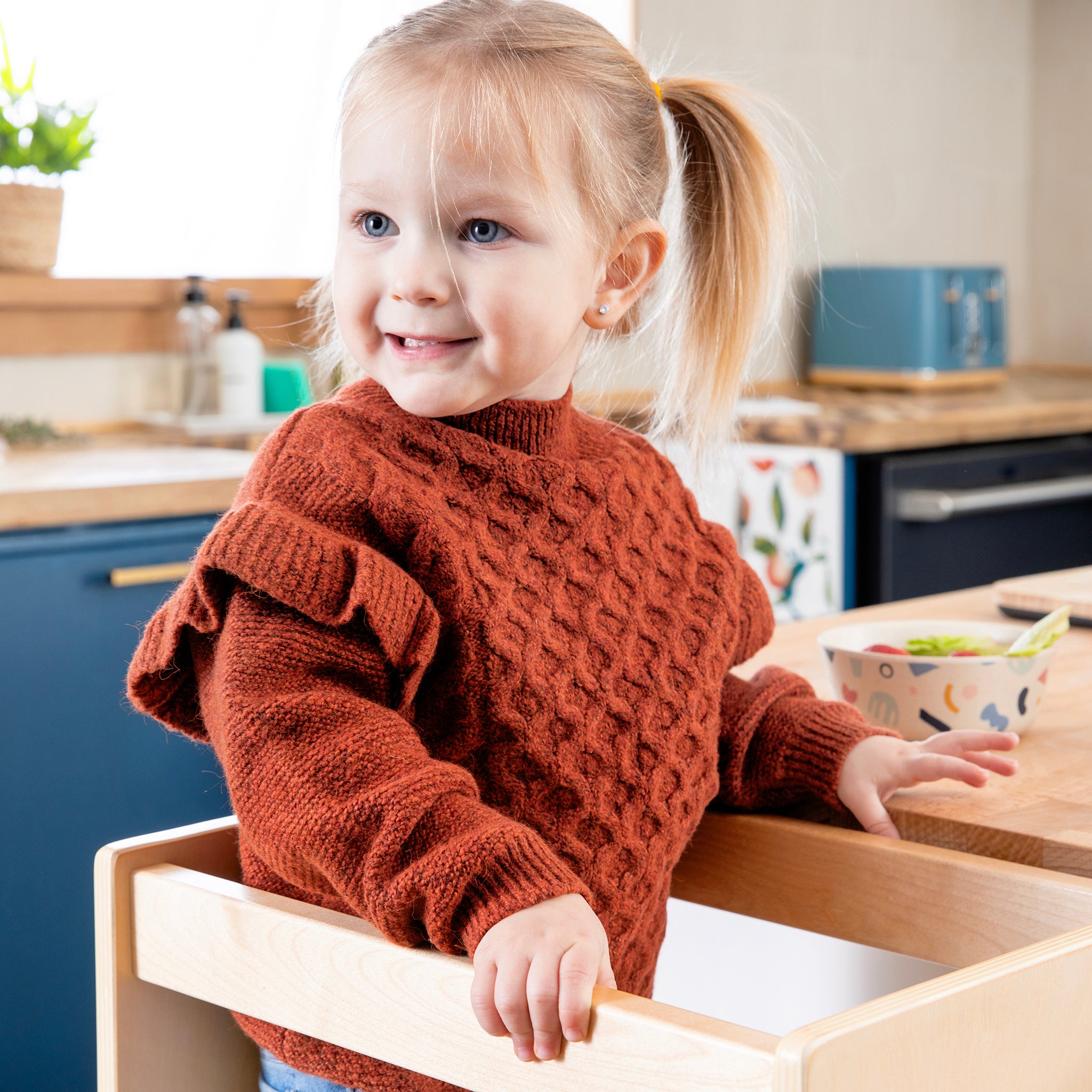 Child in a red sweater standing in a kitchen with a wooden drawer open.