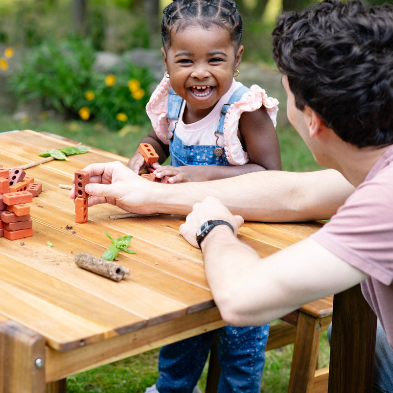 Child and adult playing with building blocks at a wooden table outdoors