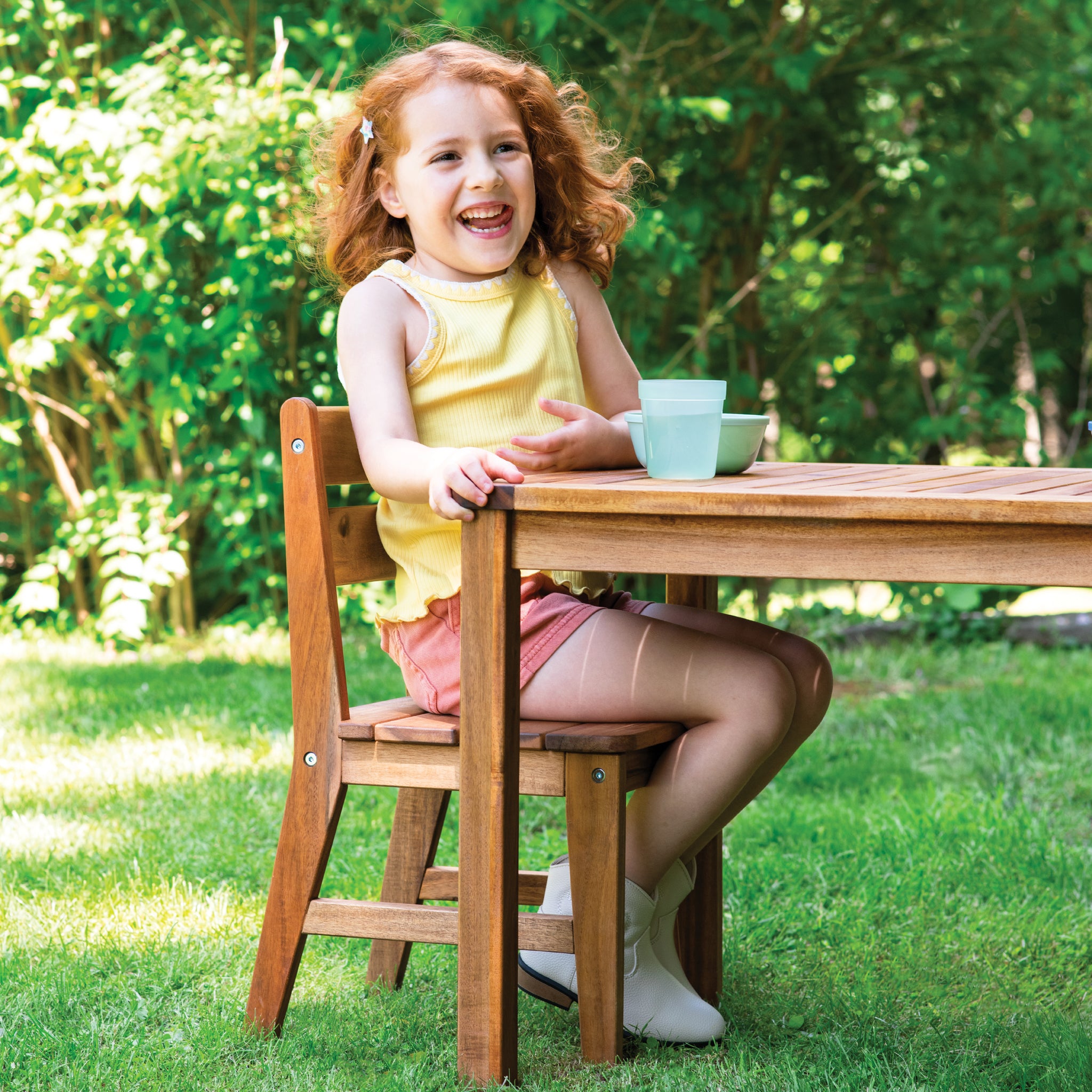 Young girl sitting at a wooden table outdoors with greenery in the background