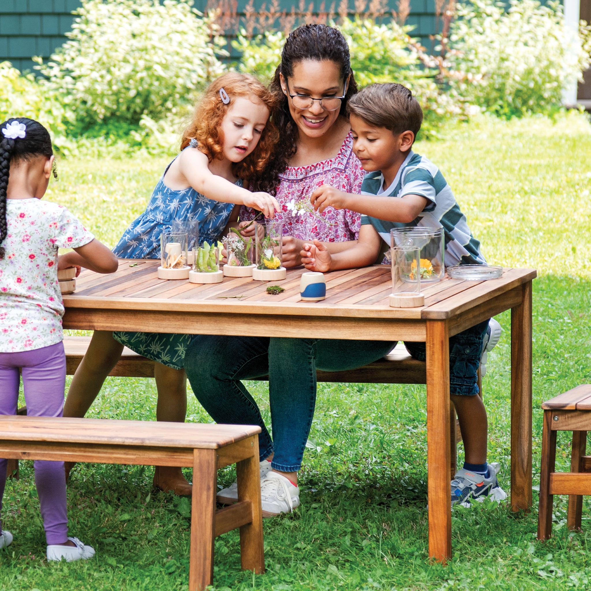 Woman and children sitting at a wooden table outdoors, engaging in an activity.