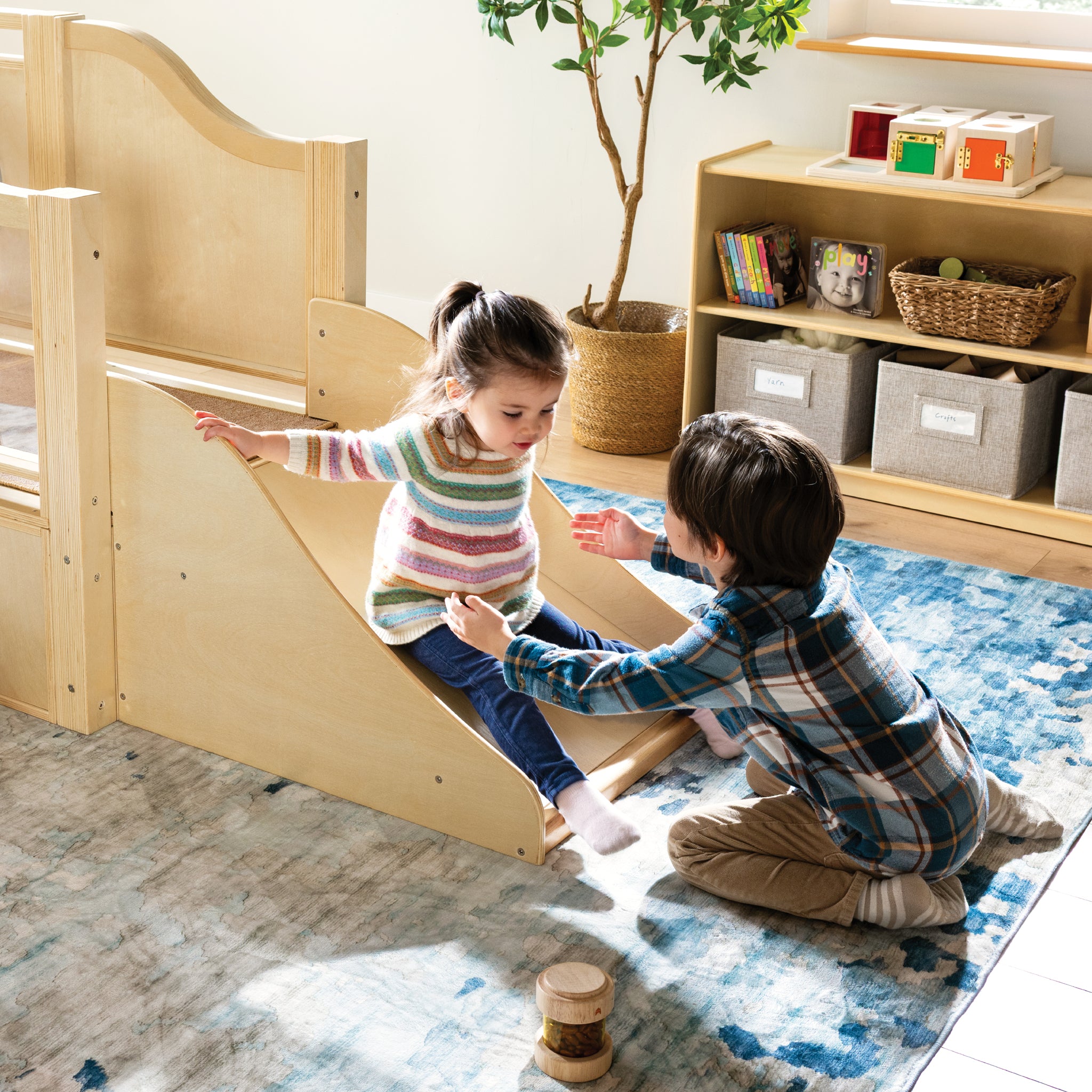 Two children playing on a wooden slide in a room with shelves and a plant.