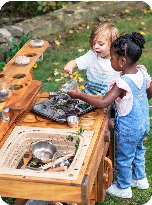 Two young children playing outdoors with a wooden play kitchen set on grass. One child pours water from a small glass pitcher into a tray, while the other stands beside the wooden counter with metal cooking utensils and dishes.