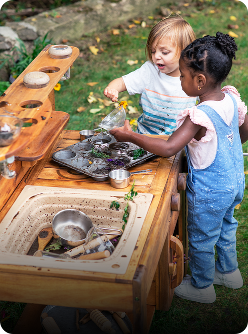 Two children playing outdoors with a wooden toy kitchen and play sink, with one child pouring from a small bottle into a tray of pretend dishes.