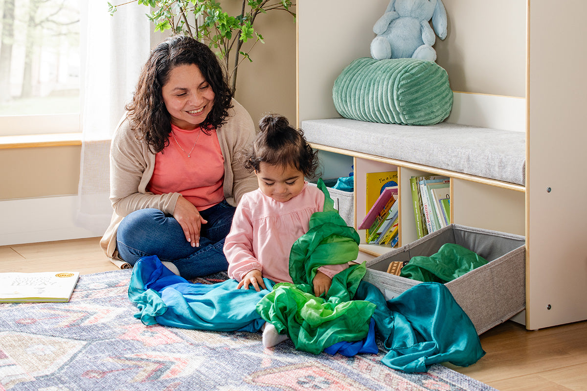Mother and child playing with green playsilks in a playroom with books and toys.