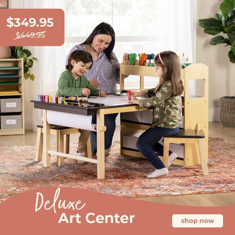Two children and a woman sitting at a wooden art center with art supplies, promotional text overlay. Click to shop the Deluxe Art Center at $349.95, save $100