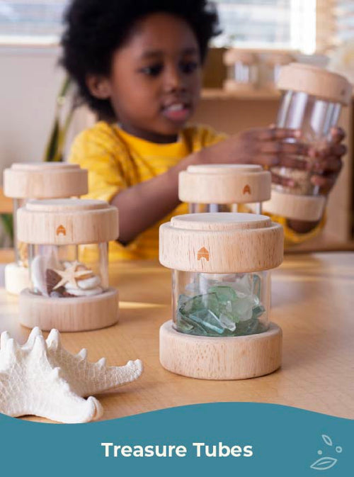 Child playing with wooden treasure tubes filled with shells and stones on a table. Click to shop Treasure Tubes