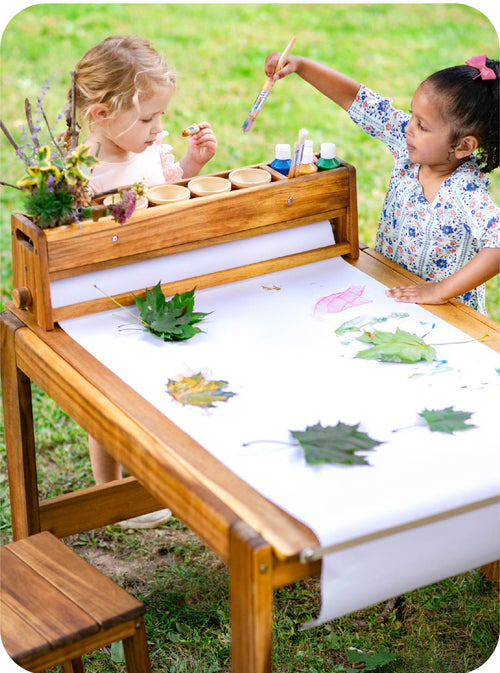 Two children at a wooden outdoor art table with nature-themed activities.